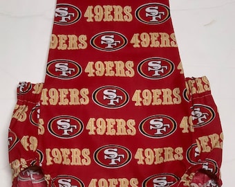 May include: Red and gold baby romper with the text "49ERS" repeated throughout. The romper has a V-neck, adjustable straps, and elasticized leg openings. The fabric is patterned with the team's logo.