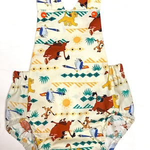 May include: A white baby onesie with a Lion King print. The onesie features Simba, Pumbaa, and Zazu, along with palm trees and a sun. The onesie has orange buttons on the straps.