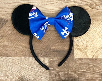 May include: Black mouse ear headband with a blue bow featuring the Los Angeles Dodgers logo and text. The ears are round and black, and the headband is black. The bow is centered between the ears.