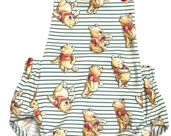 May include: A baby romper featuring a repeating pattern of a cartoon bear in various poses, wearing a red scarf. The romper has green and white striped fabric with red buttons on the straps. The garment is designed for infants and toddlers.