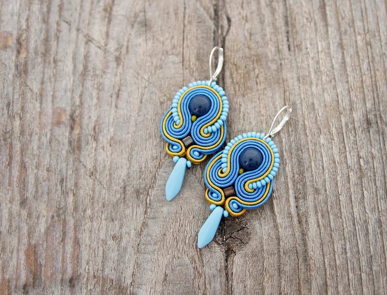 Pode incluir: Um par de brincos azuis e dourados com uma gema azul escura no centro de cada brinco. Os brincos s&atilde;o feitos de um material de cord&atilde;o tecido e t&ecirc;m um gancho prateado.