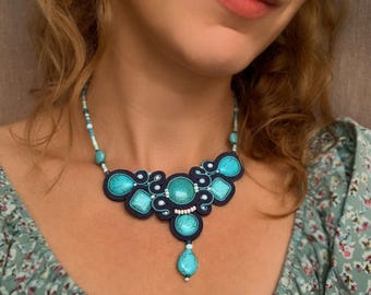 Puede incluir: Un collar de cuentas turquesa y negro con un colgante. El collar presenta una piedra turquesa grande en el centro y piedras turquesa más pequeñas alrededor. El collar está hecho de una variedad de cuentas, incluyendo cuentas turquesa, negras y blancas.