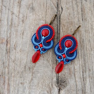 Pode incluir: Um par de brincos feitos &agrave; m&atilde;o com design soutache. Os brincos t&ecirc;m um esquema de cores vermelho e azul, com uma grande conta vermelha no centro e uma conta vermelha em forma de gota pendurada abaixo. Fundo de madeira.