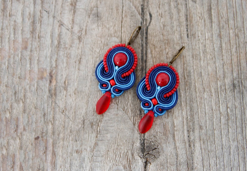Pode incluir: Um par de brincos feitos &agrave; m&atilde;o com design soutache. Os brincos t&ecirc;m um esquema de cores vermelho e azul, com uma grande conta vermelha no centro e uma conta vermelha em forma de gota pendurada abaixo. Fundo de madeira.