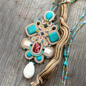 Könnte beinhalten: Eine türkisfarbene, rote und weiße Perlenkette mit einem großen, kunstvollen Anhänger. Der Anhänger ist aus geflochtenem Draht gefertigt und mit türkisfarbenen, roten und weißen Steinen besetzt.