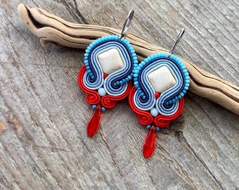 May include: A pair of red, white, and blue earrings with a white stone in the center. The earrings are made of a woven material and have a dangling drop.