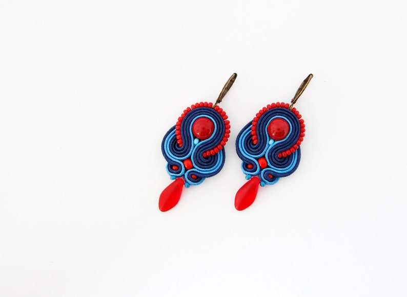 Pode incluir: Um par de brincos artesanais com design soutache. Os brincos apresentam um padr&atilde;o azul marinho e azul claro com detalhes vermelhos, incluindo uma conta central e um pingente em forma de gota. Em um fundo branco.