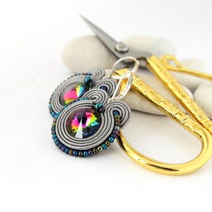 May include: A pair of silver earrings with a rainbow iridescent crystal in the center. The earrings are made with gray and silver wire and beads.