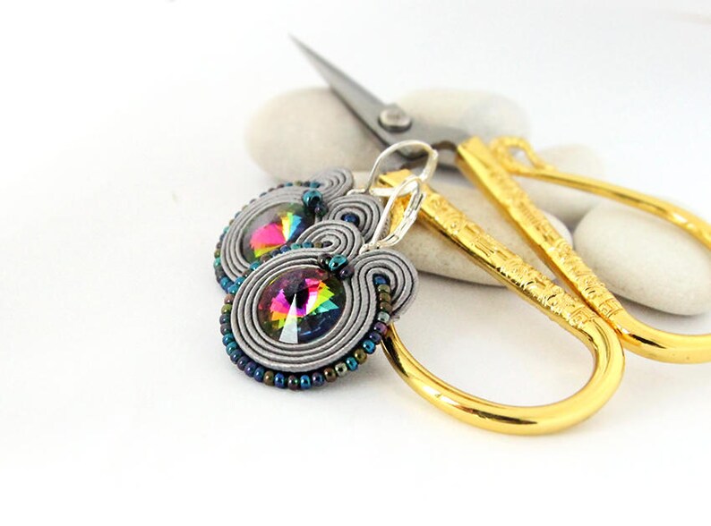 May include: A pair of silver earrings with a rainbow iridescent crystal in the center. The earrings are made with gray and silver wire and beads.