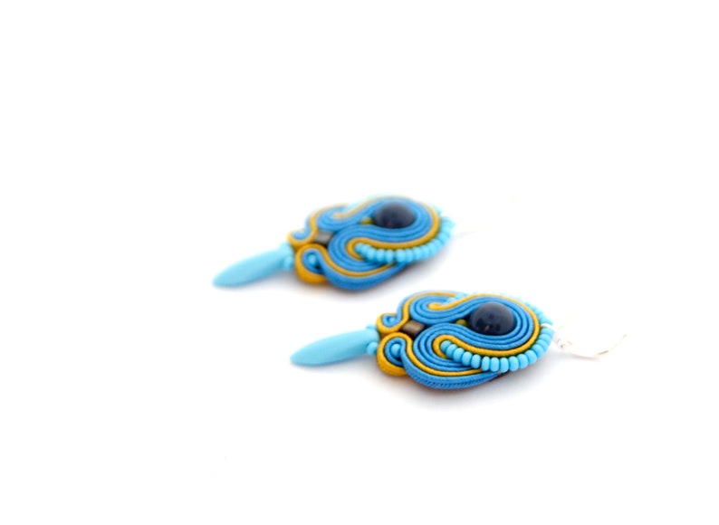 Pode incluir: Um par de brincos azuis e amarelos com uma pedra preciosa azul escura no centro. Os brincos s&atilde;o feitos de um material tecido e t&ecirc;m um gancho prateado.