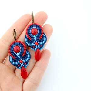 Pode incluir: Um par de brincos de soutache feitos &agrave; m&atilde;o. Os brincos apresentam uma combina&ccedil;&atilde;o de tran&ccedil;a de soutache azul marinho, azul claro e vermelho, com detalhes de contas vermelhas e pingentes vermelhos em forma de gota. Os brincos t&ecirc;m ganchos de orelha cor de bronze.