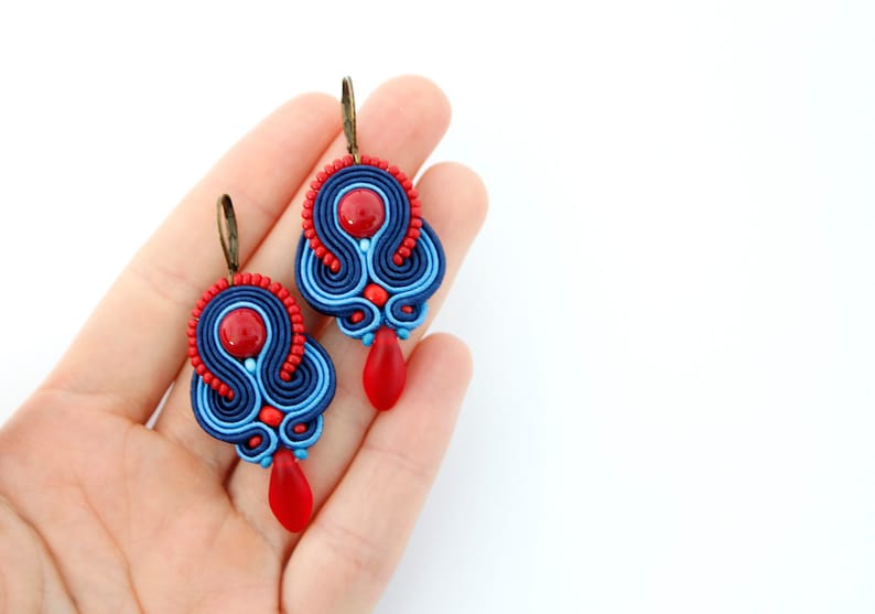 Pode incluir: Um par de brincos de soutache feitos &agrave; m&atilde;o. Os brincos apresentam uma combina&ccedil;&atilde;o de tran&ccedil;a de soutache azul marinho, azul claro e vermelho, com detalhes de contas vermelhas e pingentes vermelhos em forma de gota. Os brincos t&ecirc;m ganchos de orelha cor de bronze.