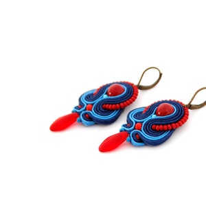Pode incluir: Um par de brincos feitos &agrave; m&atilde;o com design soutache. Os brincos apresentam uma combina&ccedil;&atilde;o de cores azul, vermelho e bronze. Cada brinco tem uma conta vermelha em forma de gota e uma conta redonda vermelha no centro.