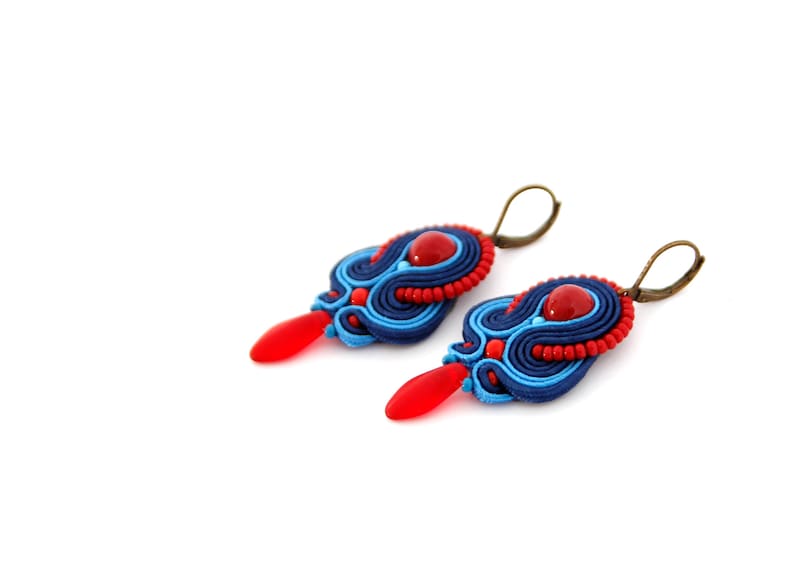 Pode incluir: Um par de brincos feitos &agrave; m&atilde;o com design soutache. Os brincos apresentam uma combina&ccedil;&atilde;o de cores azul, vermelho e bronze. Cada brinco tem uma conta vermelha em forma de gota e uma conta redonda vermelha no centro.