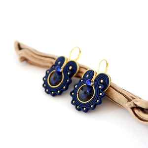 May include: A pair of blue and gold earrings with a dark blue gemstone in the center. The earrings are hanging from a wooden branch.
