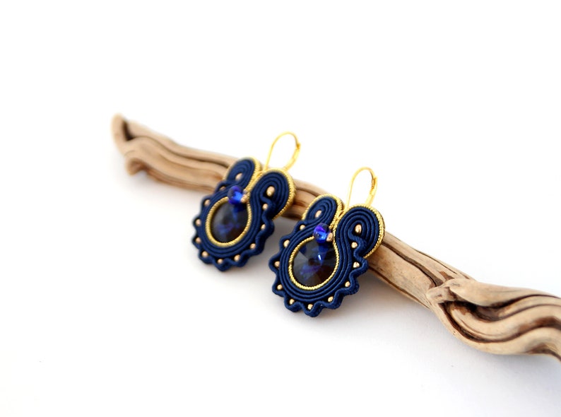 May include: A pair of blue and gold earrings with a dark blue gemstone in the center. The earrings are hanging from a wooden branch.