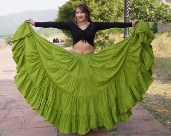 May include: A woman wearing a black long-sleeved crop top and a green, multi-layered, flowy skirt. She is standing on a brick pathway with a tree and a lamppost in the background.