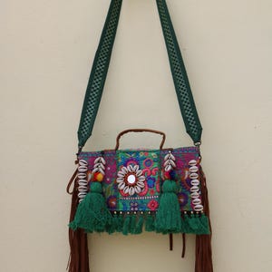 Puede incluir: Un bolso de hombro verde y marr&oacute;n con bordados coloridos, borlas y detalles de conchas. El bolso tiene un peque&ntilde;o espejo en la parte delantera y un asa de cuero marr&oacute;n.