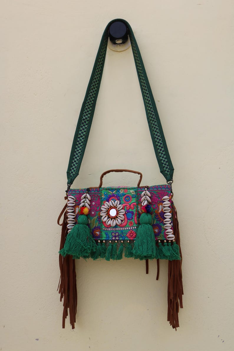 Puede incluir: Un bolso de hombro verde y marr&oacute;n con bordados coloridos, borlas y detalles de conchas. El bolso tiene un peque&ntilde;o espejo en la parte delantera y un asa de cuero marr&oacute;n.
