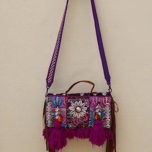 Puede incluir: Un bolso bandolera bordado en morado y rosa con flecos y detalles de conchas. El bolso tiene una correa morada con detalles dorados.