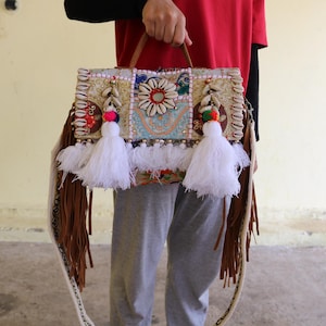 Puede incluir: Un bolso de patchwork blanco y marr&oacute;n con flecos y borlas. El bolso tiene una variedad de adornos coloridos, incluyendo conchas, cuentas y bordados.