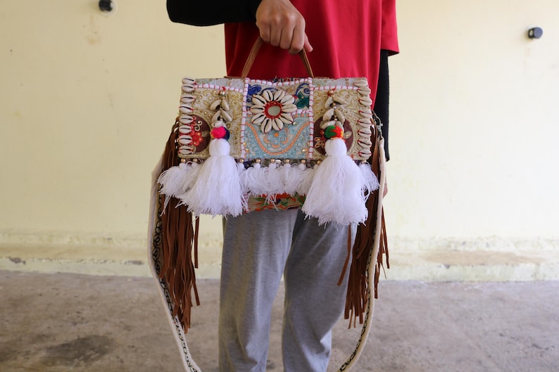 Puede incluir: Un bolso de patchwork blanco y marr&oacute;n con flecos y borlas. El bolso tiene una variedad de adornos coloridos, incluyendo conchas, cuentas y bordados.