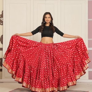 Puede incluir: Una falda roja vibrante con un estampado de lunares dorados y volantes en capas. La falda tiene un borde dorado en el dobladillo. La modelo lleva un top corto negro. La falda es una prenda festiva.