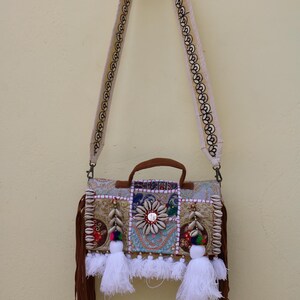 Puede incluir: Un bolso bandolera de patchwork beige y marr&oacute;n con flecos y borlas blancas. El bolso tiene una variedad de adornos, incluyendo conchas, cuentas y lentejuelas.