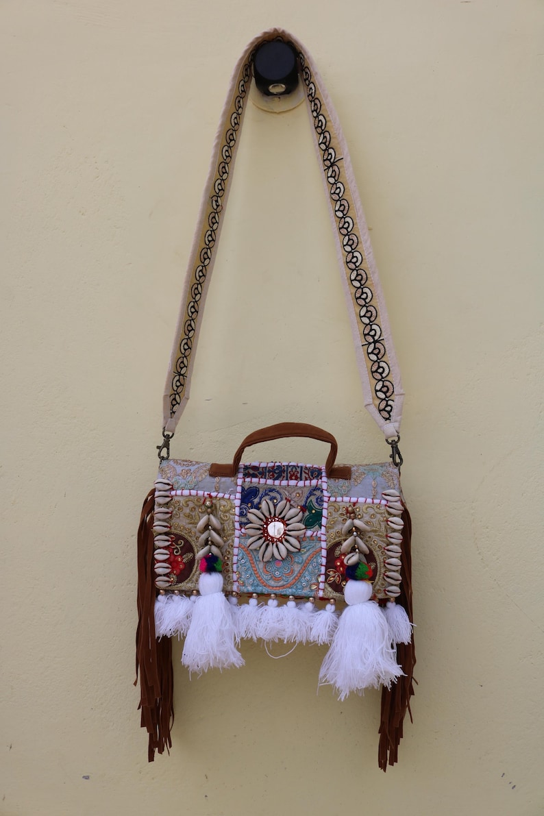 Puede incluir: Un bolso bandolera de patchwork beige y marr&oacute;n con flecos y borlas blancas. El bolso tiene una variedad de adornos, incluyendo conchas, cuentas y lentejuelas.