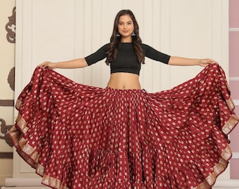 May include: A burgundy skirt with a gold pattern and trim. The skirt is tiered and flowing, with a wide waistband. The model is wearing a black crop top. The skirt is a traditional Indian garment.