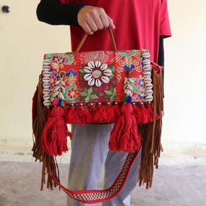 Puede incluir: Un bolso bandolera rojo, multicolor y estampado con flecos y borlas. El bolso est&aacute; decorado con conchas blancas y tiene una correa de cuero marr&oacute;n.