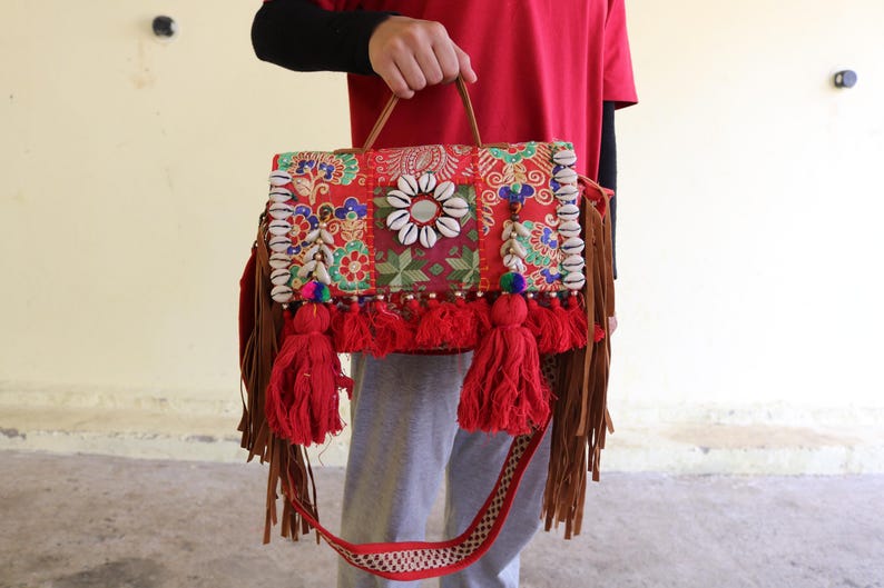 Puede incluir: Un bolso bandolera rojo, multicolor y estampado con flecos y borlas. El bolso est&aacute; decorado con conchas blancas y tiene una correa de cuero marr&oacute;n.