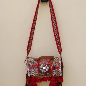 Puede incluir: Un bolso bandolera rojo de patchwork con flecos y detalles de cuentas. El bolso tiene un asa de cuero marr&oacute;n y una larga correa roja.