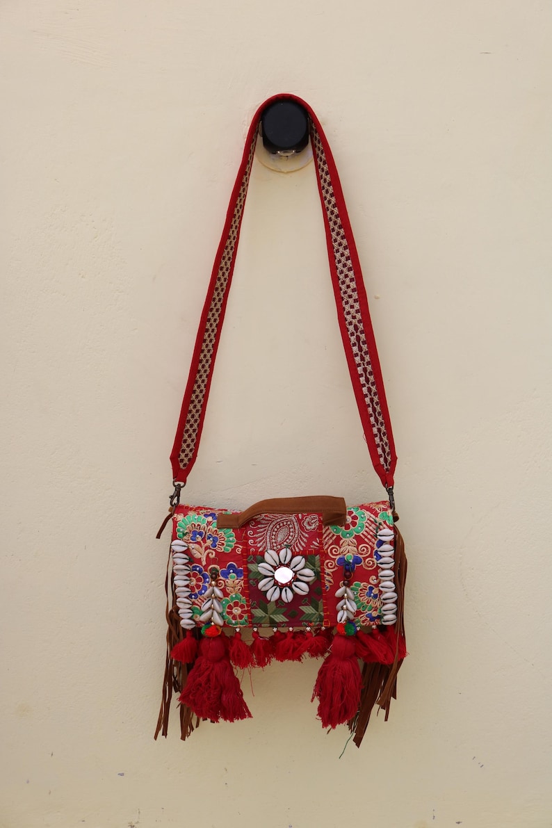 Puede incluir: Un bolso bandolera rojo de patchwork con flecos y detalles de cuentas. El bolso tiene un asa de cuero marr&oacute;n y una larga correa roja.
