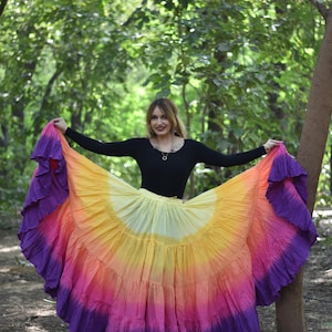 Puede incluir: Una falda multicolor con diseño escalonado, que va del amarillo al naranja, rosa y morado. La persona lleva una camiseta negra de manga larga. El fondo es un bosque verde.