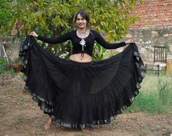 May include: A long black tiered skirt with a subtle metallic trim along the hem, paired with a black long-sleeved crop top and a silver-toned statement necklace featuring intricate metalwork and dangling elements.