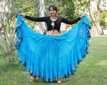 May include: A bright blue long skirt with a wide, flared silhouette and multiple horizontal tiers. The hem is finished with a decorative metallic border featuring a repeating pattern. The fabric has a subtle crinkled texture throughout the garment.