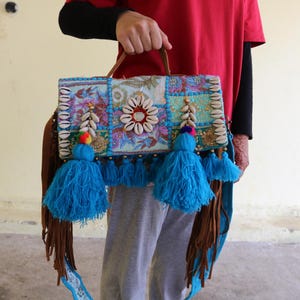 Puede incluir: Un bolso de mano de patchwork colorido con borlas azules y una correa de cuero marr&oacute;n. El bolso est&aacute; decorado con conchas marinas y cuentas.