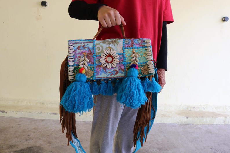 Puede incluir: Un bolso de mano de patchwork colorido con borlas azules y una correa de cuero marr&oacute;n. El bolso est&aacute; decorado con conchas marinas y cuentas.
