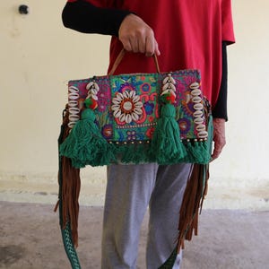 Puede incluir: Un bolso de mano de patchwork colorido con borlas verdes, detalles de conchas blancas y una correa de cuero marr&oacute;n.