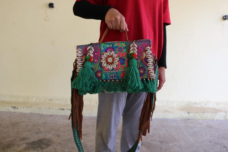 Puede incluir: Un bolso de mano de patchwork colorido con borlas verdes, detalles de conchas blancas y una correa de cuero marr&oacute;n.