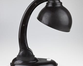 May include: A dark brown desk lamp with a curved arm and a rounded, dome-shaped shade. The lamp features a solid oval base with a small white push-button switch located on the top surface. The structure is smooth and uniform in color.