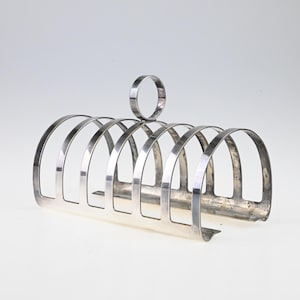 May include: A silver-toned metal toast rack featuring six arched slots for holding slices of bread. The structure is composed of a horizontal base with vertical curved dividers and a single circular handle centered at the top for easy carrying.