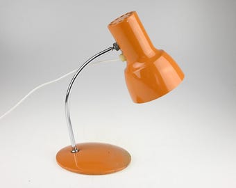 May include: A bright orange desk lamp featuring a cylindrical head with a perforated top, a curved chrome metal arm, and a circular orange base. A white power cord extends from the back of the lamp head, which is adjustable via a small hinge.