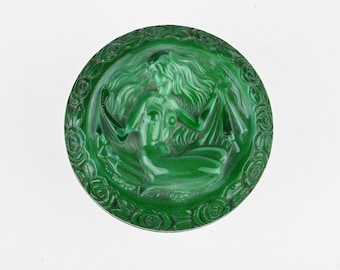 May include: A circular green glass decorative plate featuring a central relief carving of a reclining figure with long flowing hair. The outer border of the plate is decorated with a repeating pattern of sculpted roses in a matching green tone.