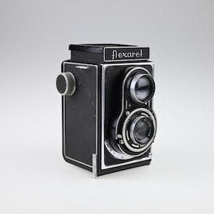 May include: A vintage black Flexaret twin-lens reflex camera with silver trim. The camera has two lenses, a top viewfinder lens and a bottom taking lens. The word "flexaret" is printed on the top of the camera.