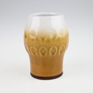 May include: A white and brown ceramic drinking glass with a textured design. The glass has a rounded bottom and a wide mouth.
