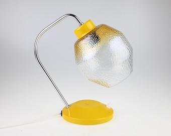 May include: A table lamp featuring a bright yellow circular base and a matching yellow socket housing. A curved chrome metal arm supports a textured, clear glass shade that transitions into a subtle amber gradient at the top and bottom edges.