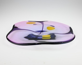 May include: A shallow, organic-shaped glass bowl featuring a translucent light purple base with deep violet accents. The surface is decorated with three distinct, solid yellow circular spots and dark blue glass swirls embedded within the center.