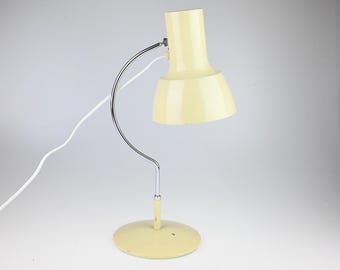 May include: A cream-colored desk lamp featuring a curved chrome metal arm, a circular base, and a cylindrical shade. The lamp includes a white power cord extending from the back of the shade, providing a functional lighting solution for a workspace.